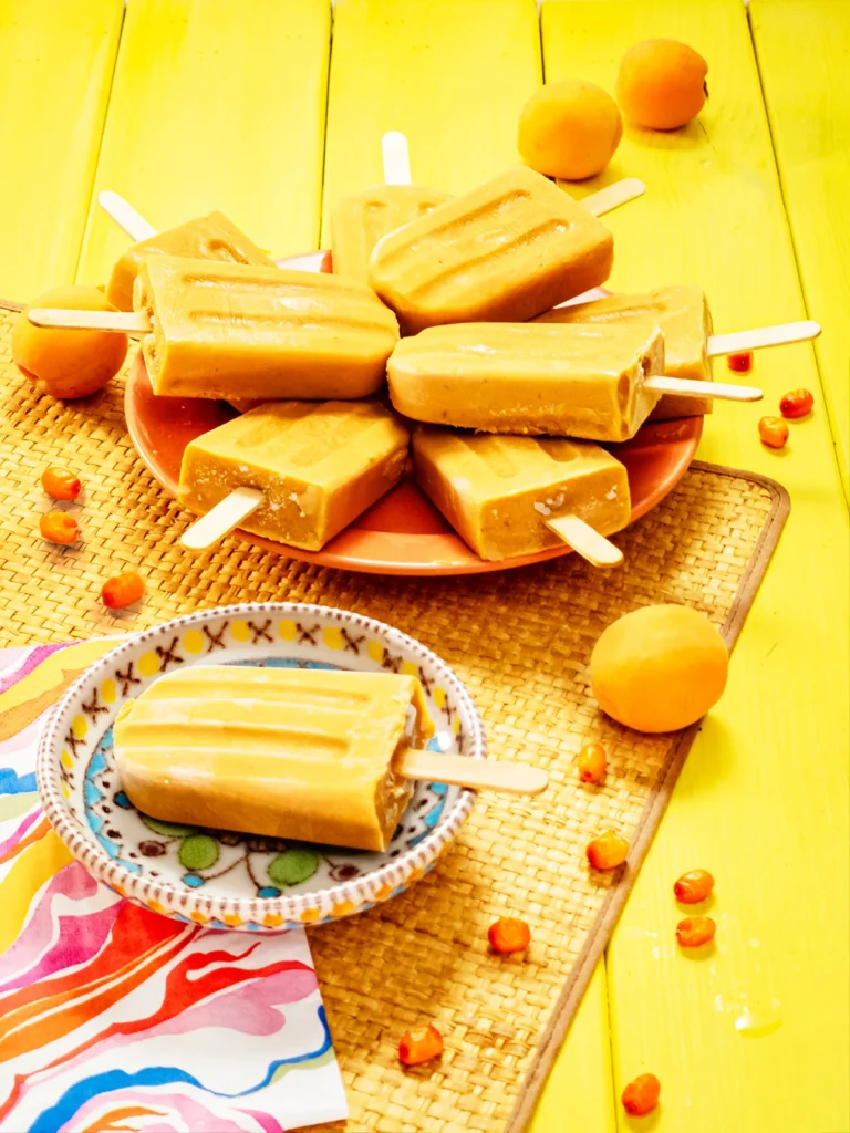 There is a sea buckthorn popsicle on a small colourful plate. On the left is a patterned napkin. Behind the small plate is an orange serving plate with a lots of sea buckthorn popsicles. The plates are placed on a grass mat. Apricots and sea buckthorn berries are scattered on the yellow wooden base and the mat.