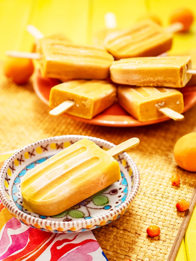 There is a sea buckthorn popsicle on a small colourful plate. On the left is a patterned napkin. With a blurred view, behind the small plate is an orange serving plate with a lots of sea buckthorn popsicles. The plates are placed on a grass mat. Apricots and sea buckthorn berries are scattered on the yellow wooden base and the mat.