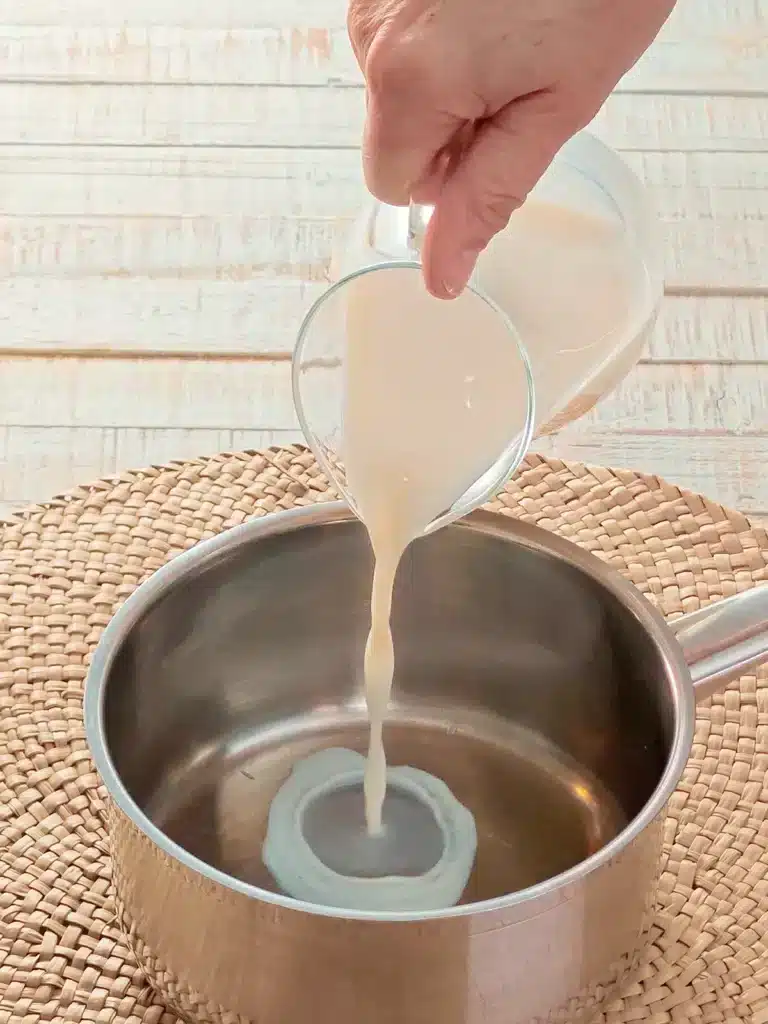 A hand pours oat milk from a glass jug into a stainless steel pot standing on a seaweed mat.