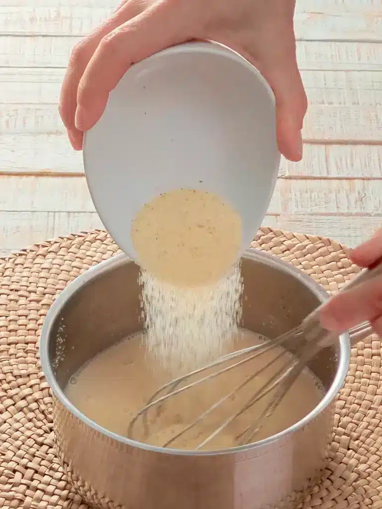 A hand pours semolina into a stainless steel pot of warm oat milk. The pot stands on a seaweed mat. In the background is a white wooden base.