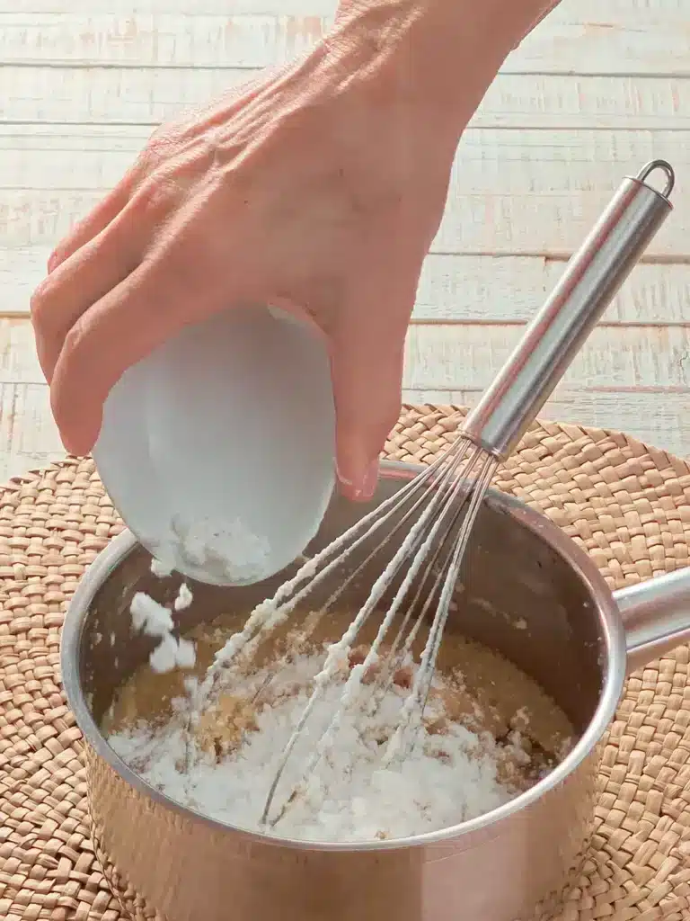 A hand pours potato starch into a stainless steel pot with a whisk. The pot stands on a seaweed mat. In the background is a white wooden base.