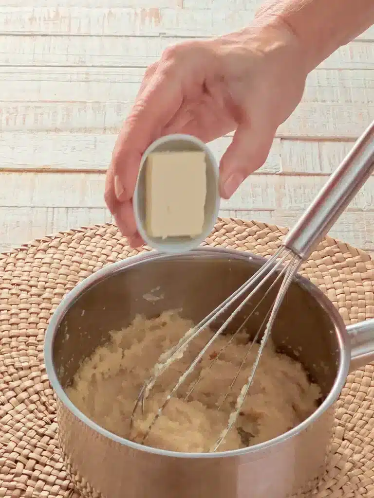 A hand pours vegan butter into a stainless steel pot with mash and whisk. The pot stands on a seaweed mat. In the background is a white wooden background.