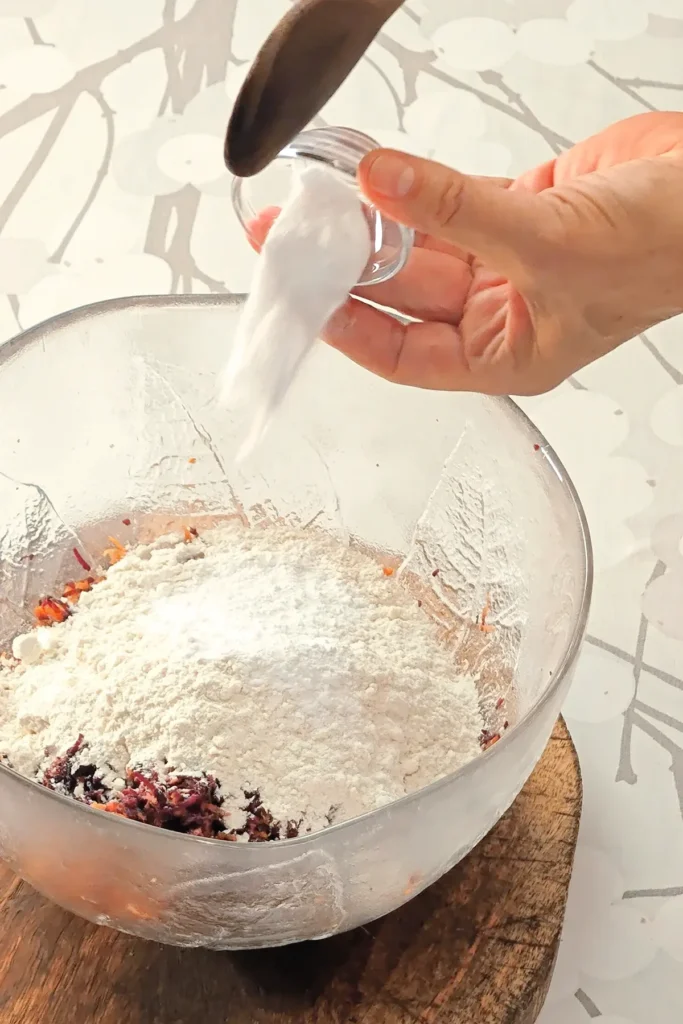 A hand is holding a small glas bowl with baking soda, which is pured into the glass bowl with the ingredients for the base cake of the Hummus Hazelnut Cake. The bowl stands on a wooden coaster on a tablecloth with a branched pattern.