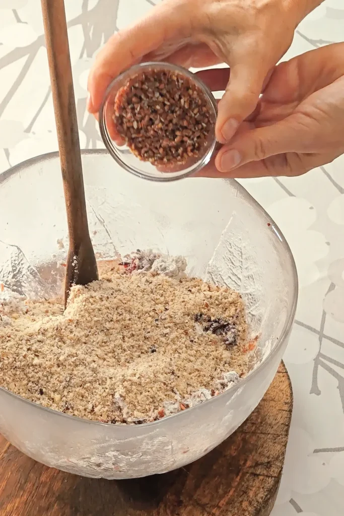 A hand is holding a small glass bowl with flaxseed eggs, which is pured into the glass bowl with the ingredients for the base cake of the Hummus Hazelnut Cake. The bowl stands on a wooden coaster on a tablecloth with a branched pattern.