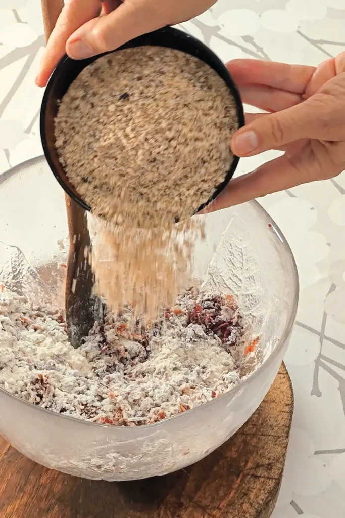 A hand is holding a bowl with grounded hazelnuts, which is pured into the glass bowl with the ingredients for the base cake for the Hummus Hazelnut Cake. The bowl stands on a wooden coaster on a tablecloth with a branched pattern.