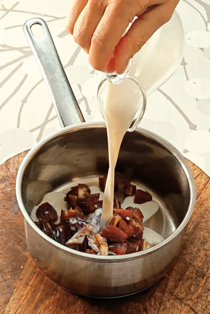 One hand holds a small jug of oat milk, which is poured into a steel pot with the chopped dates. The pot stands on a wooden trivet on a tablecloth with a branched pattern.