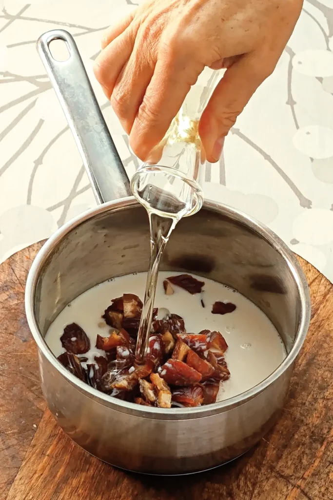 One hand holds a glass of oil, which is poured into the date and oat milk mixture in a steel pot. The pot stands on a wooden trivet on a tablecloth with a branched pattern.