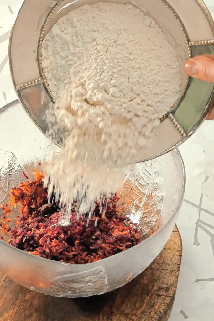 A hand is holding a bowl with flour, which is pured into the glass bowl with the ingredients for the base cake of the Hummus Hazelnut Cake. The bowl stands on a wooden coaster on a tablecloth with a branched pattern.