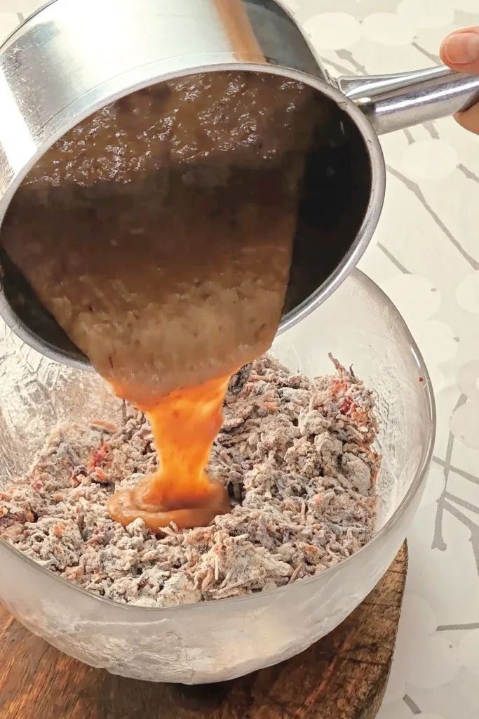 A hand is holding a stainless steel pot with the purred mixture of dates, which is pured into the glass bowl with the ingredients for the base cake of the Hummus Hazelnut Cake. The bowl stands on a wooden coaster on a tablecloth with a branched pattern.