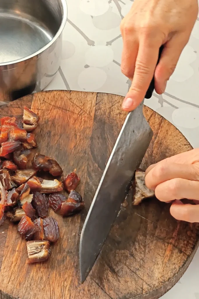 A hand is holding a huge knife, that is cutting the dates on a wooden cutting board. The wooden board is placed on a tablecloth with a branched pattern. In the background ther is a stainless steel pot.