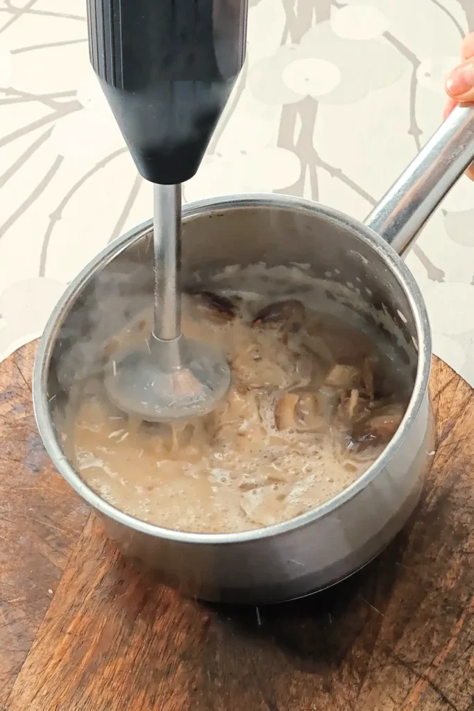 A hand blender is making a purred mixture of dates, oat milk, and oil, for the Base of the Hummus Hazelnut Cake. All in a stainless steel pot that stands on a wooden plate on a tablecloth with a branch pattern.