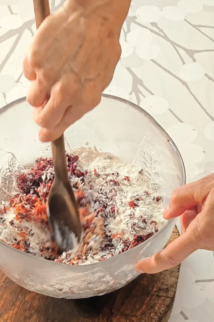 A hand is holding a glass bowl that stands on a wooden coaster on a tablecloth with a branched pattern. The other hand is holding a wooden spoon and stir together the ingredients for the base cake of the Hummus Hazelnut Cake.