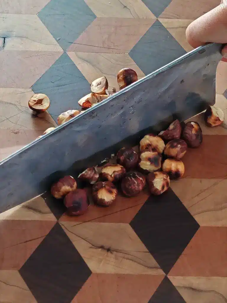 One hand holds a large knife that chops up roasted hazelnuts. The nuts lie on a patterned wooden board.