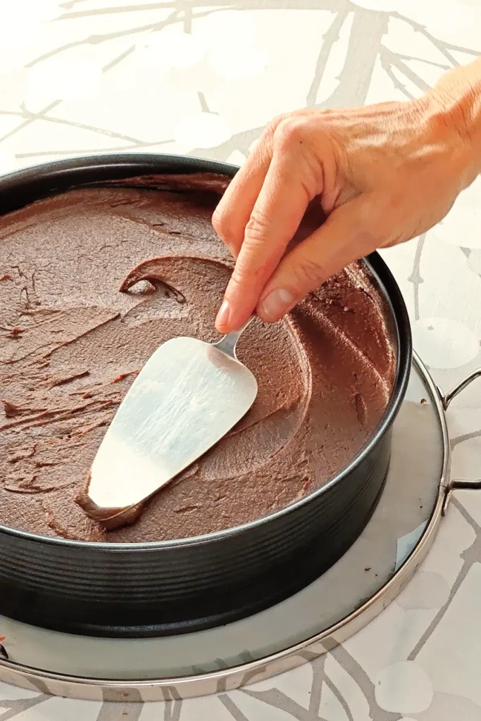 One hand spreads the hummus cocoa cream on the cake base of the Hummus Hazelnut Cake, with the help of a cake lifter. The springform pan is placed on a cake serving tray on a tablecloth with a branched pattern.