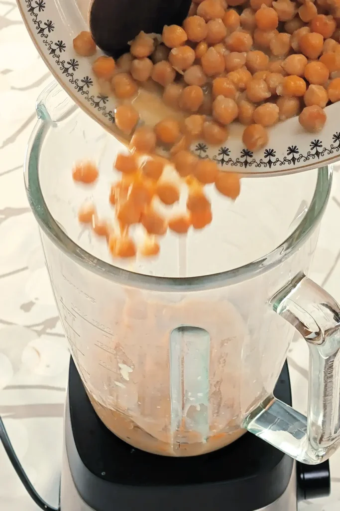 A blender, already filled with a mixture of ingredients for the cream of the Hummus Hazelnut Cake, is placed on tablecloth with a branch pattern. Two hands hold a bowl with chickpeas, it is filled into the blender with the help of a wooden spoon.