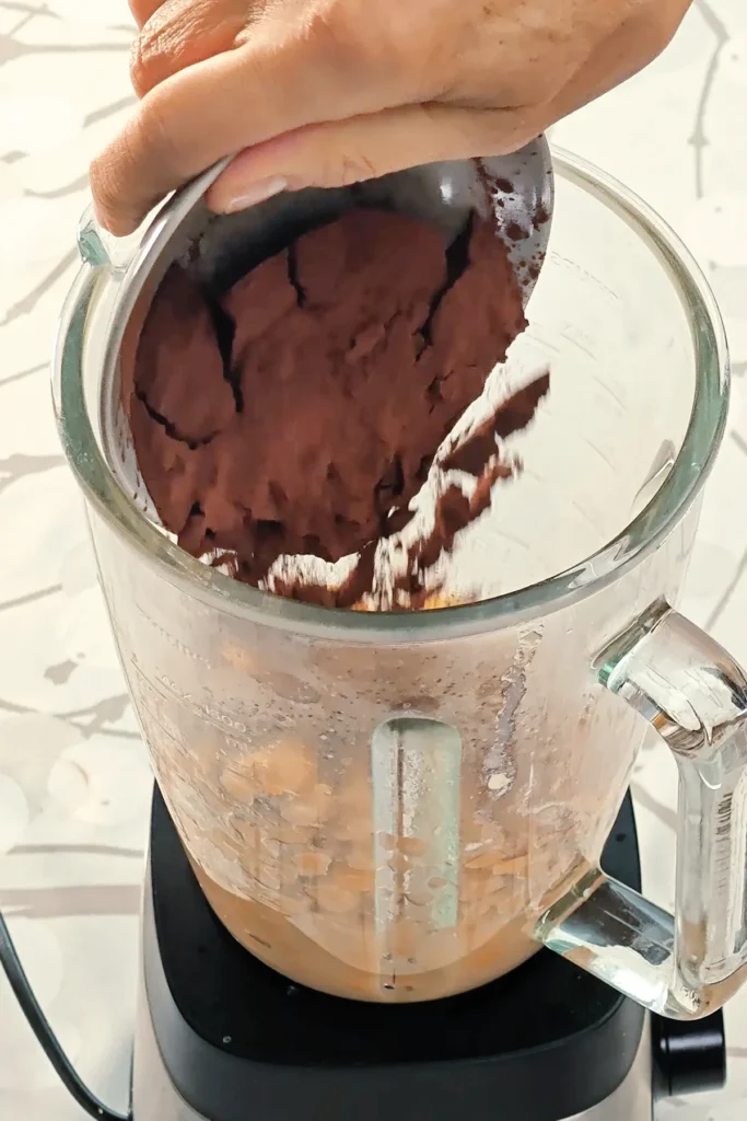 A blender, already filled with a mixture of ingredients for the cream of the Hummus Hazelnut Cake, is placed on tablecloth with a branch pattern. A hand holds a bowl with cocoa powder, it is filled into the blender.