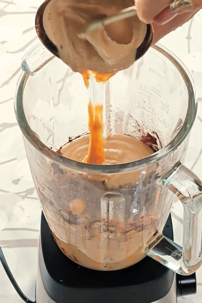 A blender, already filled with a mixture of ingredients for the creamof the Hummus Hazelnut Cake, is placed on tablecloth with a branch pattern. Two hands hold a bowl with tahini, it is filled into the blender with the help of a spoon.