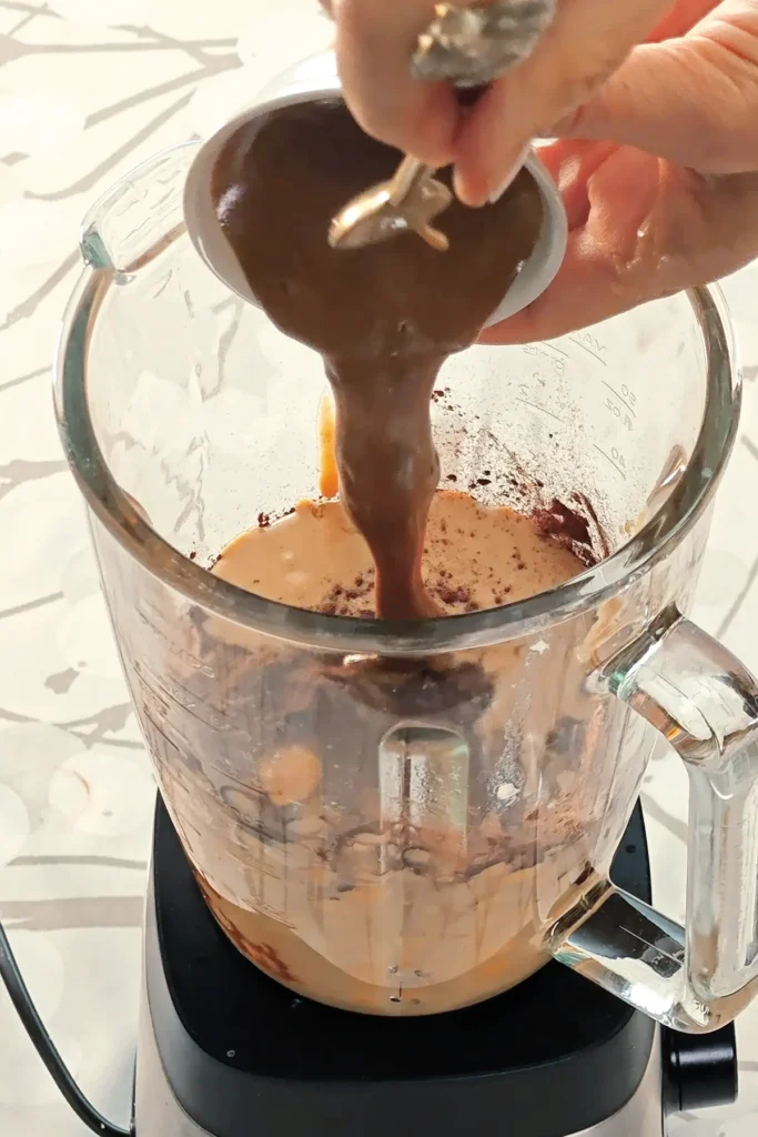 A blender, already filled with a mixture of ingredients for the cream of the Hummus Hazelnut Cake, is placed on tablecloth with a branch pattern. Two hands hold a bowl with hazelnut puree, it is filled into the blender with the help of a spoon.