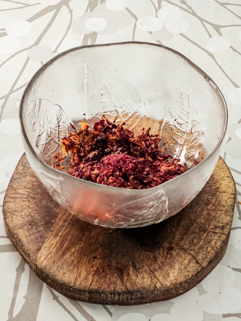 A glass bowl stands on a wooden coaster on a tablecloth with a branched pattern. There are grated carrots in the bowl.