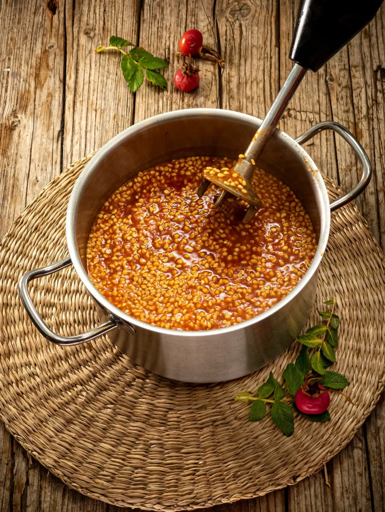 A raffia mat lies on a wooden base, with a stainless steel pot on top. In the pot is the Rose Hip Soup with lots of seeds floating on the surface. In the pot is a hand blender with which the soup has just been pureed. Some rose hip twigs and rose hip fruits lie around the pot.