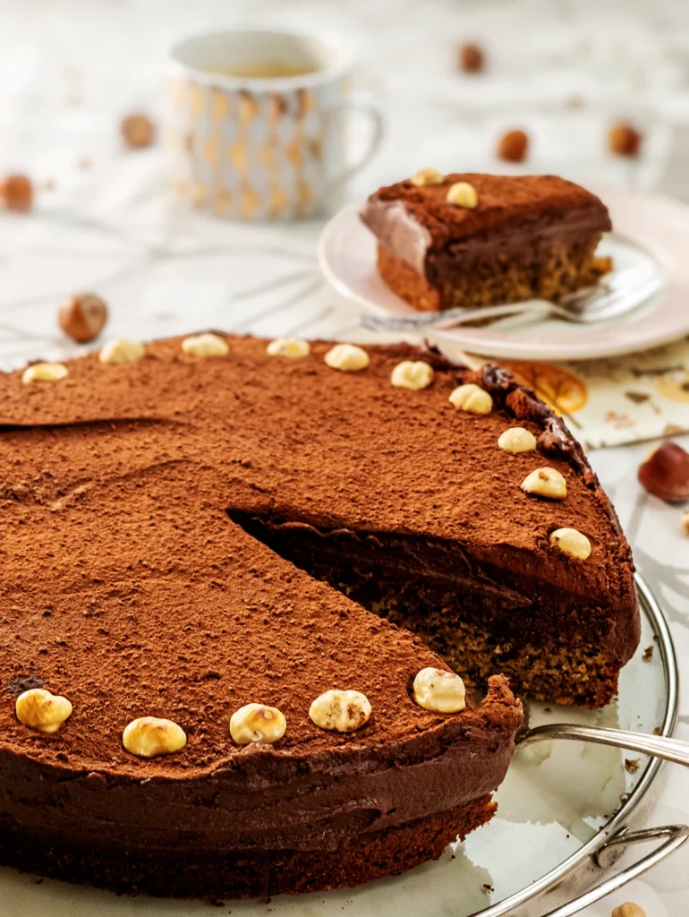 In the foreground, is a cake serving plate with the Hummus Hazelnut Cake with a silver cake server. Behind on the right side, slightly blurred, a piece of the Hummus Hazelnut Cake is served on a plate with a silver cake fork next to it. Under the plate, you can see a patterned napkin. On the left side stands a cup of coffee with a golden pattern print. All are placed on a table cloth with a branch pattern on it. Hazelnuts and dried chickpeas are scattered around.