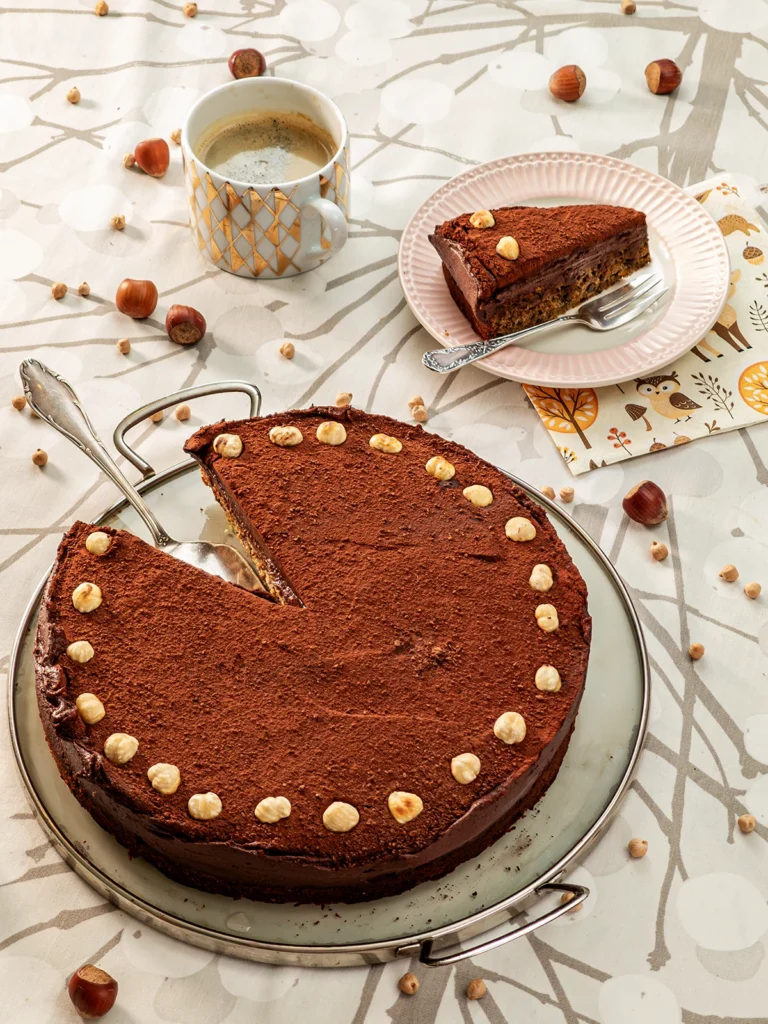 In the foreground, is a cake serving plate with the Hummus Hazelnut Cake, with a silver cake server. Behind on the right side, a piece of the Hummus Hazelnut Cake is served on a plate with a silver cake fork next to it. Under the plate, you can see a patterned napkin. On the left side stands a cup of coffee with a golden pattern print. All are placed on a table cloth with a branch pattern on it. Hazelnuts and dried chickpeas are scattered around.