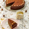 In the foreground, a piece of the Hummus Hazelnut Cake is served on a plate with a silver cake fork next to it. Under the plate, you can see a patterned napkin. On the right side stands a cup of coffee with a golden pattern print. Behind is a cake serving plate with the Hummus Haselnut Cake with a silver cake server. All are placed on a table cloth with a branch pattern on it. Hazelnuts and dried chickpeas are scattered around.