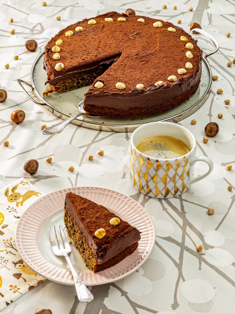 In the foreground, a piece of the Hummus Hazelnut Cake is served on a plate with a silver cake fork next to it. Under the plate, you can see a patterned napkin. On the right side stands a cup of coffee with a golden pattern print. Behind is a cake serving plate with the Hummus cake with hazelnut with a silver cake server. All are placed on a table cloth with a branch pattern on it. Hazelnuts and dried chickpeas are scattered around.