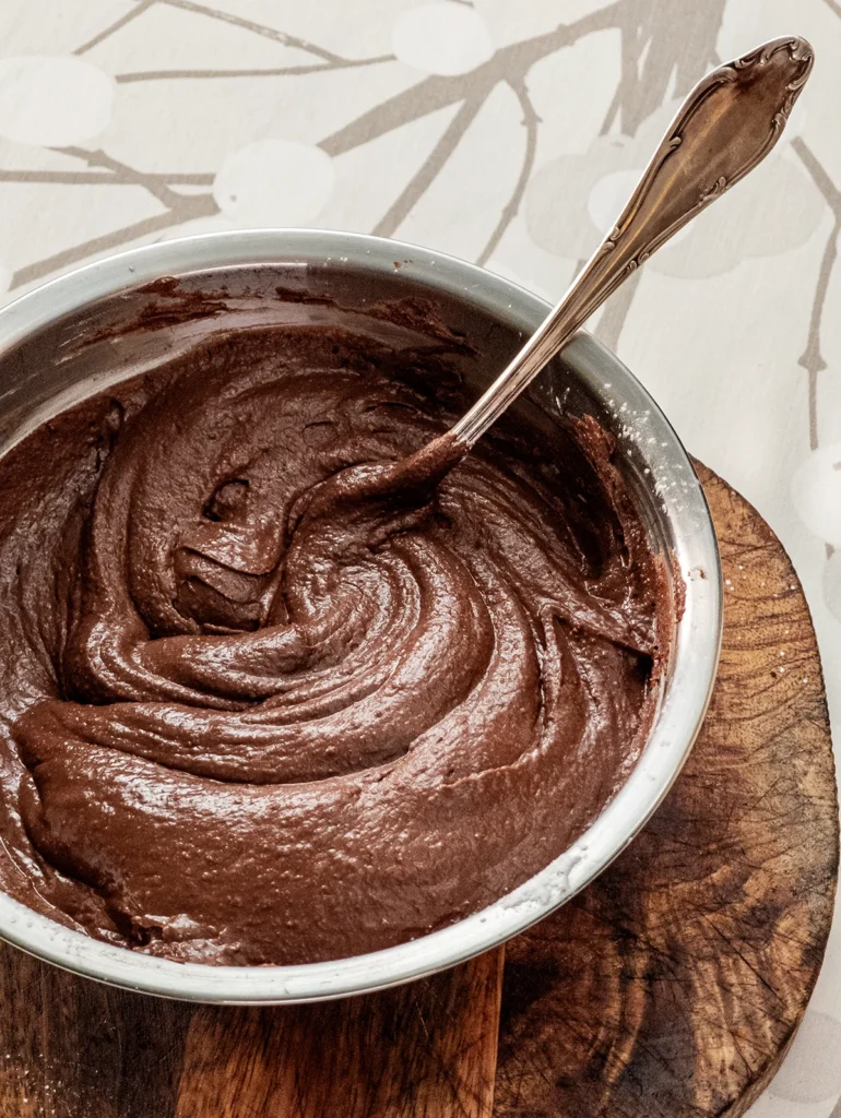 A stainless steel bowl with a silver fork filled with hummus cacao cream stands on a wooden plate on a tablecloth with a branch pattern.