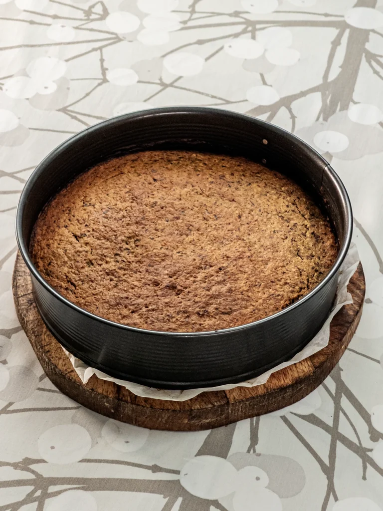 The springform pan with the baked hazelnut cake base stands on a wooden plate on a tablecloth with a branch pattern.