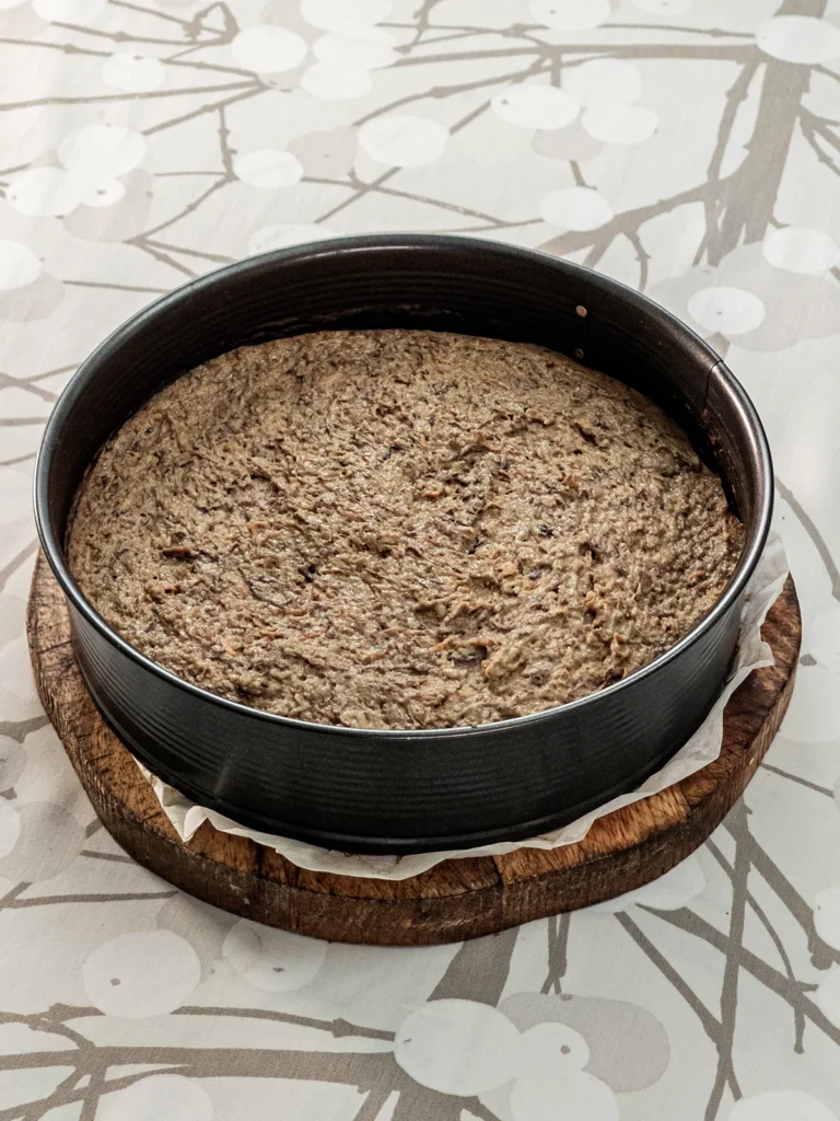 The springform pan with the dough for the Hummus Hazelnut Cake. It stands on a wooden plate on a tablecloth with a branch pattern.