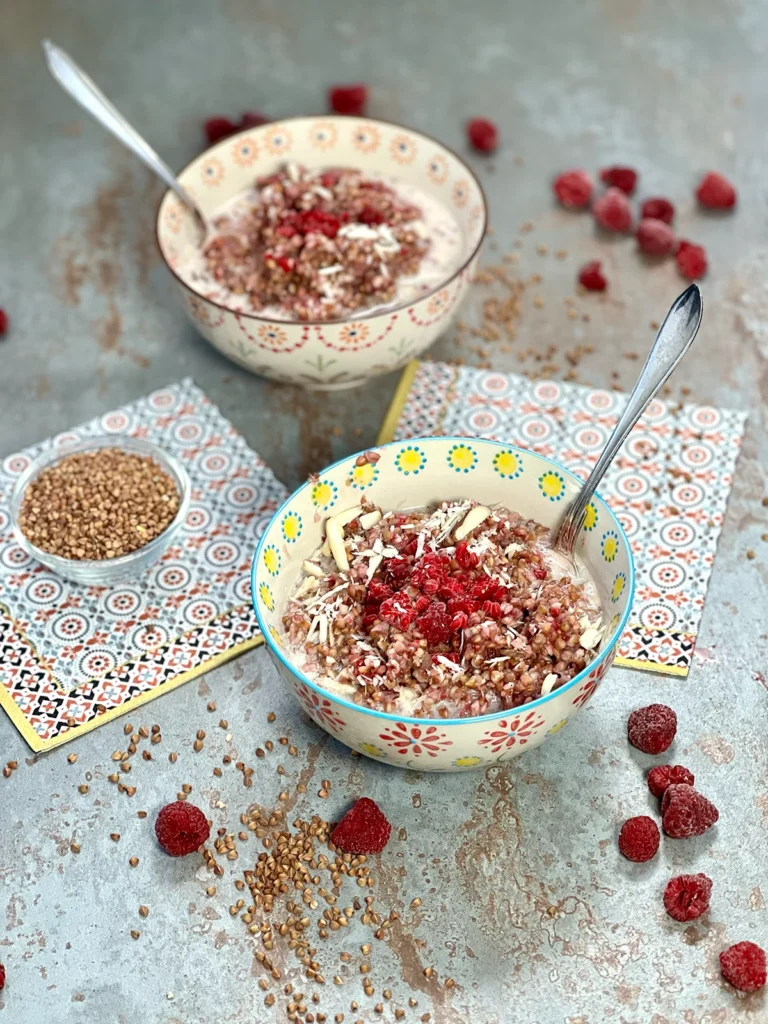 Two beautiful bowls of buckwheat porridge, garnished and with frozen raspberries around them