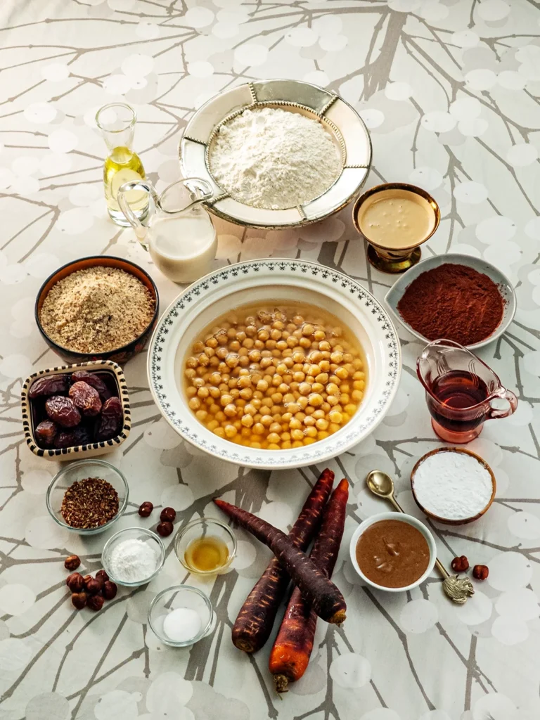 Here you see all the ingredients to make a Hummus Hazelnut Cake.There are many bowls on a tablecloth with a branch pattern. In the large bowl in the center are chickpeas in aquafaber. Around it are bowls of flour, tahini, cocoa for baking, starch, hazelnut puree, grounded flaxseeds, Medjool dates, and grounded hazelnuts. Between the bowls, you can also see glass jars with oat milk, maple syrup, and oil. In the foreground are three smaller glass bowls with apple cider vinegar, baking soda, and baking powder. Next to them are three fresh carrots. Some hazelnuts are scattered around, and a decorative spoon with a squirrel lies next to the bowl with hazelnut puree.