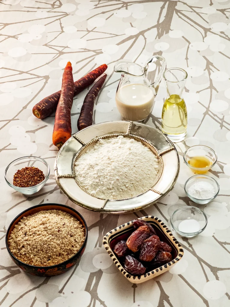 Here you can see all the ingredients for the base to make a Hummus Hazelnut Cake. There are seven bowls of different sizes on a tablecloth with a branch pattern. In the large bowl in the middle is flour. Around the flour were bowls of Medjool dates, grounded flaxseeds, and grounded hazelnuts. There are three smaller glass bowls with apple cider vinegar, baking soda, and baking powder. Behind the bowls, you can see two glass jars with oat milk and oil and three fresh carrots.