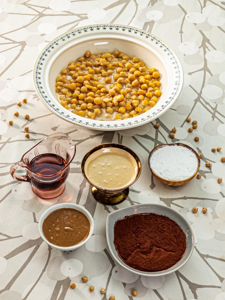 Here you can see all the ingredients for the cream on the Hummus Hazelnut Cake. There are five bowls on a tablecloth with a branch pattern. In the large bowl in the background are chickpeas in aquafaber. Before, there were bowls of tahini, starch, hazelnut puree, and cocoa for baking. There is also a jar with maple syrup. Some dried chickpeas are scattered around.