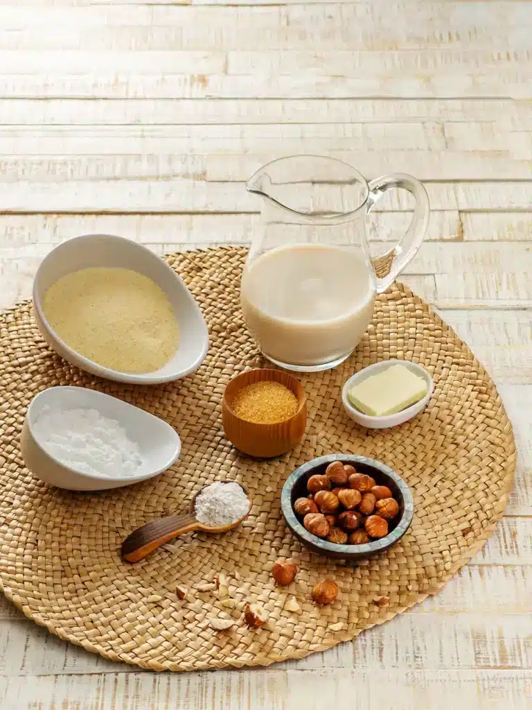 Bowls of semolina, potato starch, cane sugar, vegan butter and whole hazelnuts are placed on a round seaweed mat. There is also a jug of oat milk. In the foreground lies a wooden spoon with vanilla sugar and some hazelnut pieces are scattered around. The seaweed mat lies on a white wooden base.