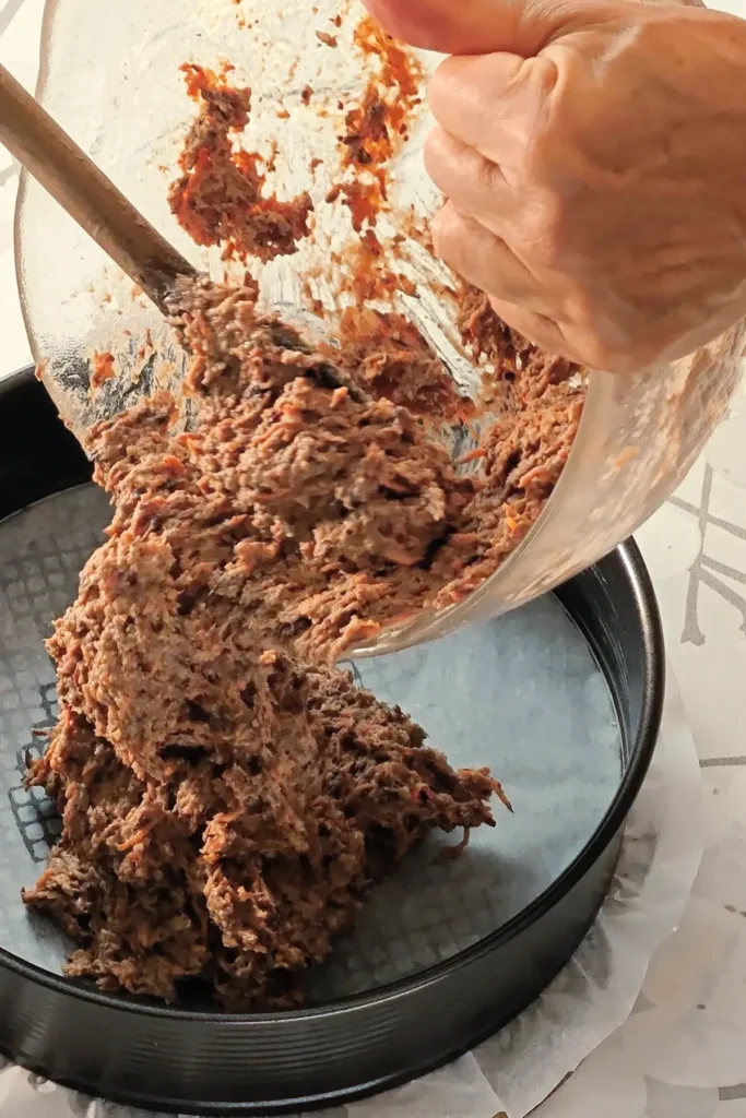 The hazelnut carrot pastry for the base of the Hummus Hazelnut Cake, is spread in the springform tin. The springform pan is placed on a wooden board on a tablecloth with a twig pattern.