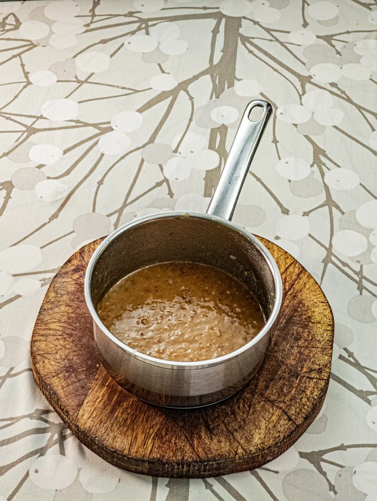 A stainless steel pot with the purred mixture with the dates, stands on a wooden plate on a tablecloth with a branch pattern. The mixture is needed for making the cake base of the Hummus Hazelnut Cake.