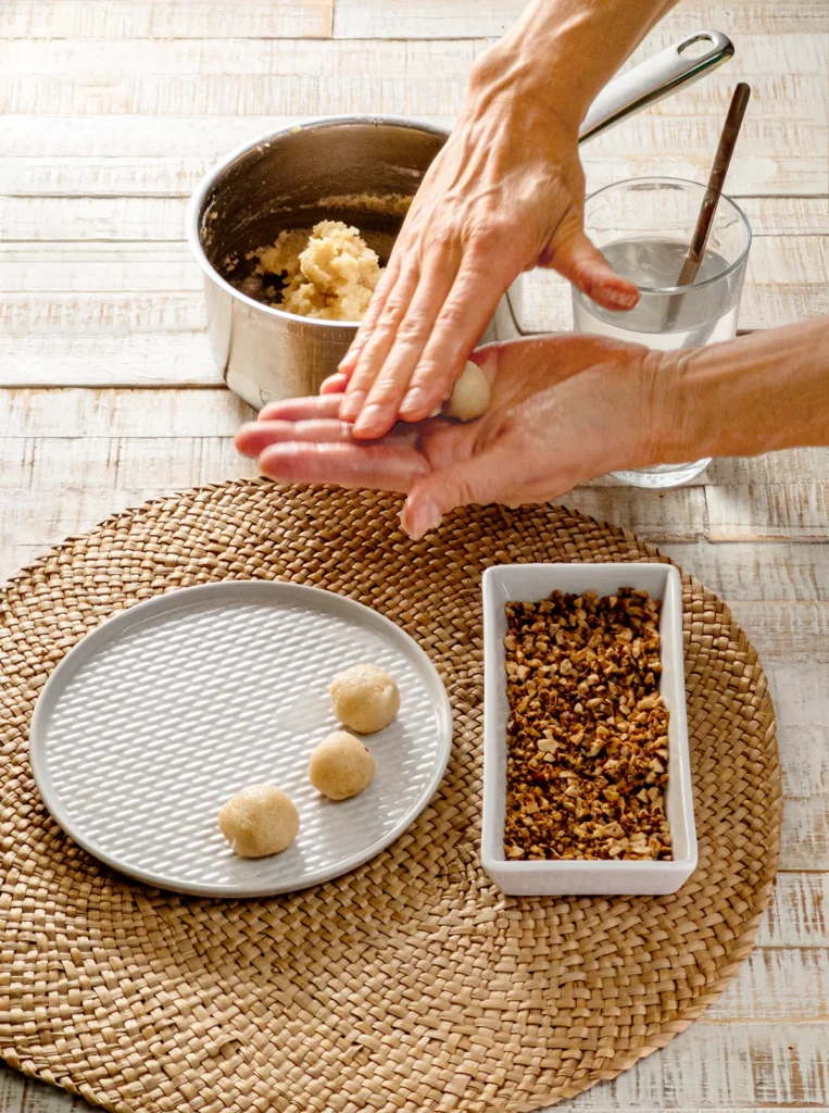 On a round seaweed mat there are a white plate and a square shallow bowl. There are chopped roasted hazelnuts in the bowl and three semolina dumplings on the plate. Behind the mat is a stainless steel pot of semolina porridge. Next to it, is a glass of water with a long spoon. Two hands roll a ball out of the porridge. The seaweed mat lies on a white wooden base.