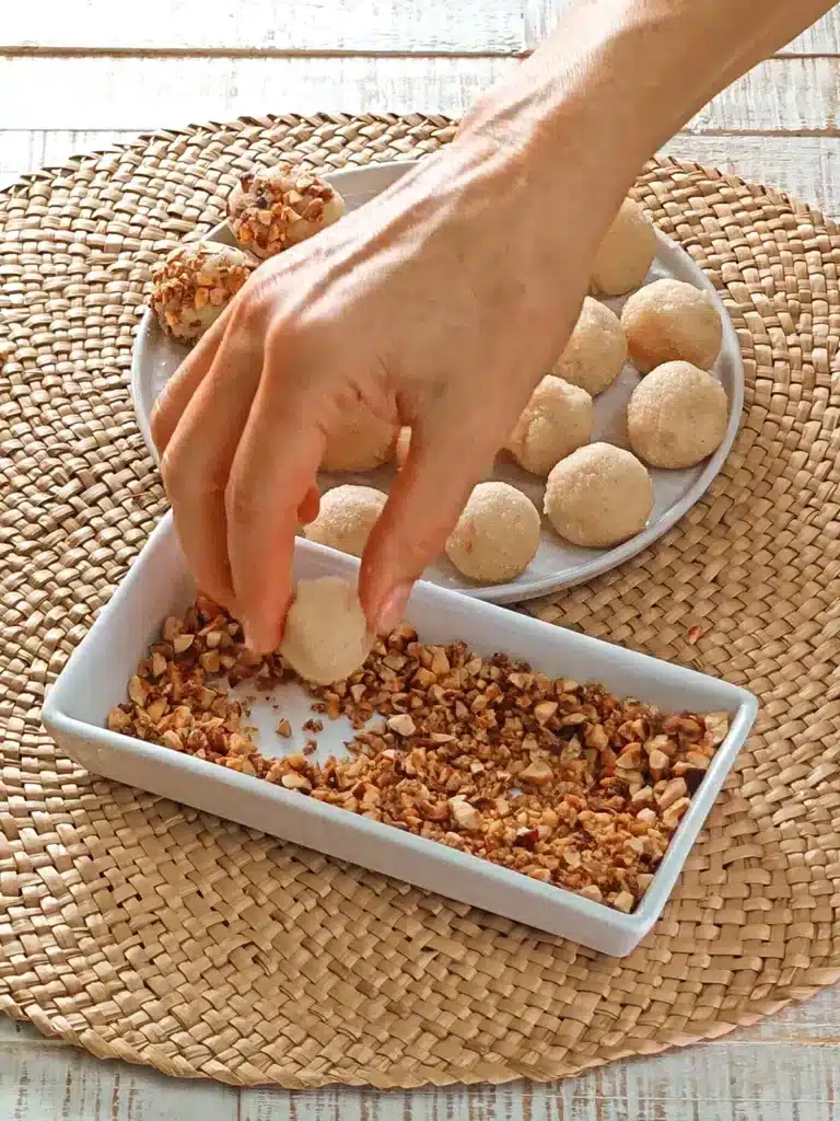 On a round seaweed mat there are a white plate and a square shallow bowl. There are chopped roasted hazelnuts in the bowl and semolina dumplings on the plate. One hand holds a semolina dumpling and adds it to the chopped hazelnuts. The seaweed mat lies on a white wooden base.