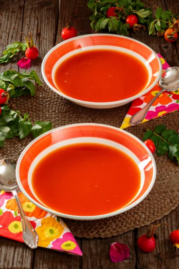 On a wooden base, there is a raffia mat on which two soup plates are placed. The plates are filled with Rose Hip Soup. Next to the plates are napkins with a floral pattern and a silver soup spoon. Rose hip twigs and rose hip fruits lie around the soup plates.