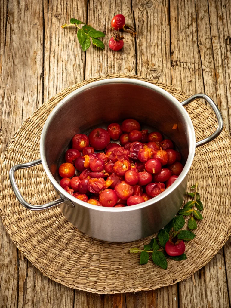 A raffia mat lies on a wooden base, with a stainless steel pot on top. In the pot are the washed and de-stemmed rose hip fruits for making Rose Hip Soup. Some rose hip twigs and rose hip fruits lie around the pot.