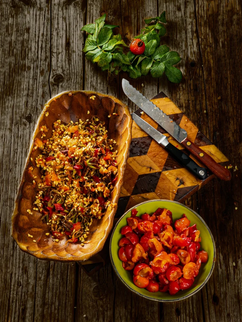 A patterned wooden cutting board stands on a wooden base, on top is an elongated wooden bowl full of rosehip seeds and flower clippings. Next to it are the knives used, a short kitchen knife and a larger serrated knife. In the foreground is a green bowl filled with ready-pitted rosehip fruits. In the background are some rosehip twigs with a rosehip fruit.