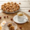 A seagrass mat lies on a white wooden base. In the foreground is a light gray bowl with two semolina balls with roasted hazelnuts. Half under the bowl is a patterned napkin. Behind it on the right is a white demitasse cup with a saucer and a silver spoon, filled with coffee. In the background is a light gray plate with more Semolina dumplings with roasted hazelnuts. A few hazelnuts are scattered around the place setting.