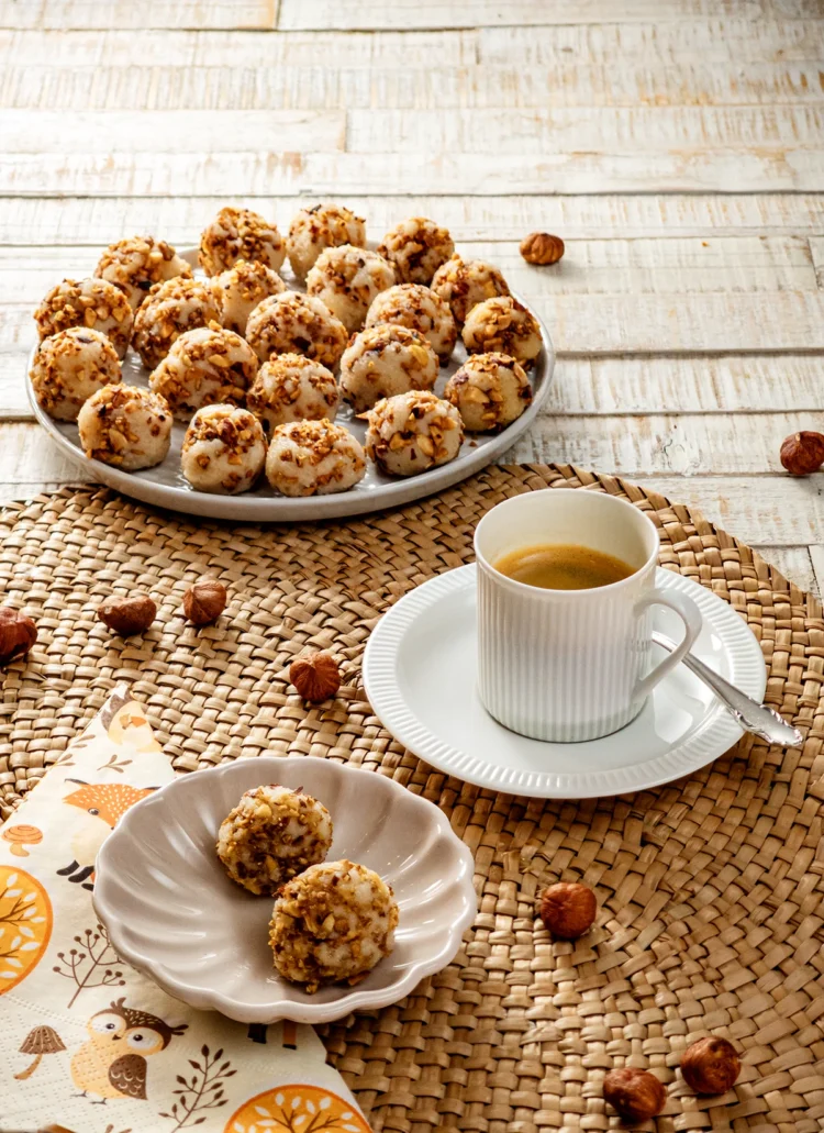 A seagrass mat lies on a white wooden base. In the foreground is a light gray bowl with two semolina balls with roasted hazelnuts. Half under the bowl is a patterned napkin. Behind it on the right is a white demitasse cup with a saucer and a silver spoon, filled with coffee. In the background is a light gray plate with more Semolina dumplings with roasted hazelnuts. A few hazelnuts are scattered around the place setting.