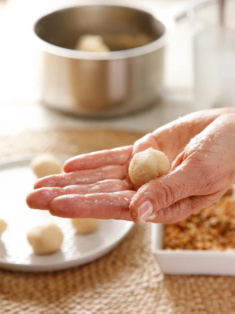 On a seaweed mat there are a white plate and a square shallow bowl. There are chopped roasted hazelnuts in the bowl and semolina dumplings on the plate. In the background is a pot. One hand holds a semolina dumpling in the foreground.