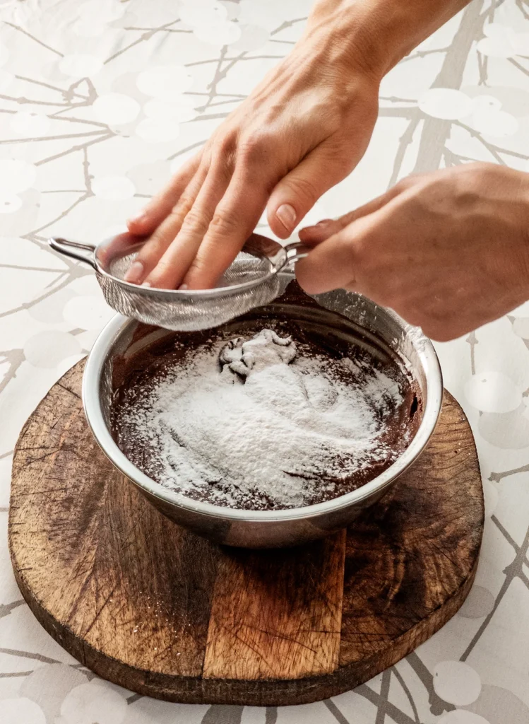 One hand rubs the starch into the hummus and cocoa cream. A stainless steel bowl filled with hummus and cocoa cream is placed on a wooden plate on a tablecloth with a branch pattern.