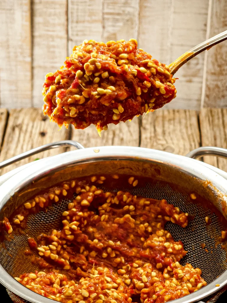 A pot with a sieve can partly be seen on a wooden base. In the sieve, that is on the pot, you can see the rest of the cooked rose hip fruits, namely lots of seeds. A silver spoon also shows a mountain of seeds and leftover fruit.