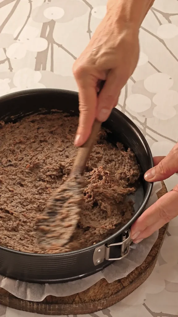 The hazelnut carrot pastry for the base of the Hummus Hazelnut Cake, is spread in the springform tin. The springform pan is placed on a wooden board on a tablecloth with a branch pattern.