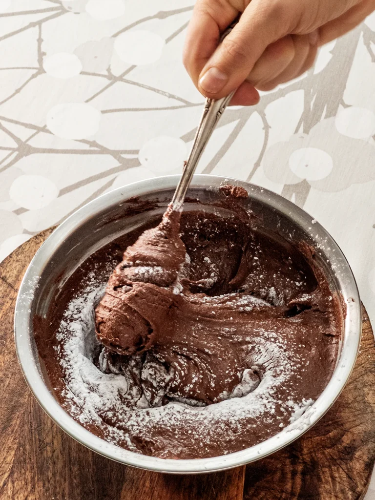 One hand stirs the starch into the hummus cacao cream with a silver fork to thicken it. The hummus cacao cream is in a stainless steel bowl, placed on a wooden plate on a tablecloth with a branch pattern.